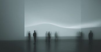 Ghostly silhouettes of people moving below an abstract light in a dark room.